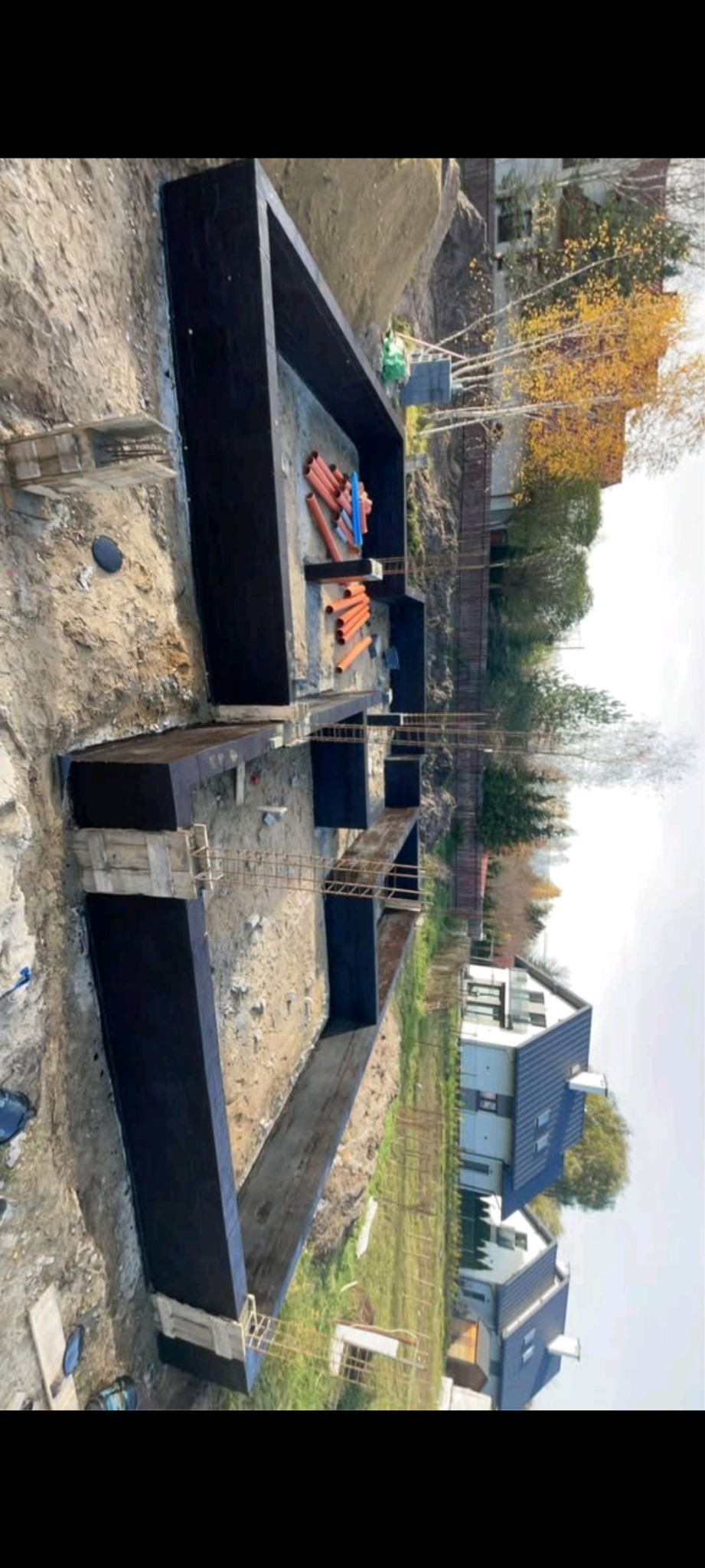 Wykopane fundamenty domu z hydroizolacją, widoczne szalunki i zbrojenie, na działce leżą rury kanalizacyjne, w tle domy sąsiadów.