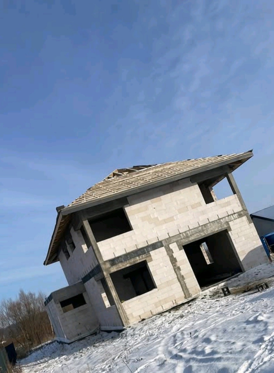 Dwukondygnacyjny dom w stanie surowym otwartym, zbudowany z białych bloczków, widoczny na tle zimowego krajobrazu i błękitnego nieba.