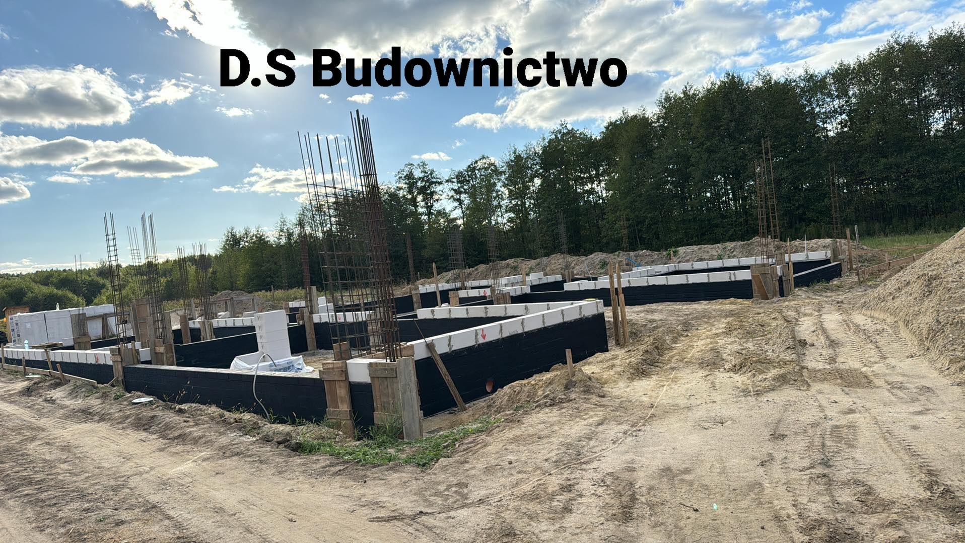 Fundamenty domu w trakcie budowy, zbrojenie pionowe, izolacja przeciwwilgociowa, teren budowy z piaskiem i widocznym lasem w tle, logo firmy D.S Budownictwo nałożone na zdjęcie.
