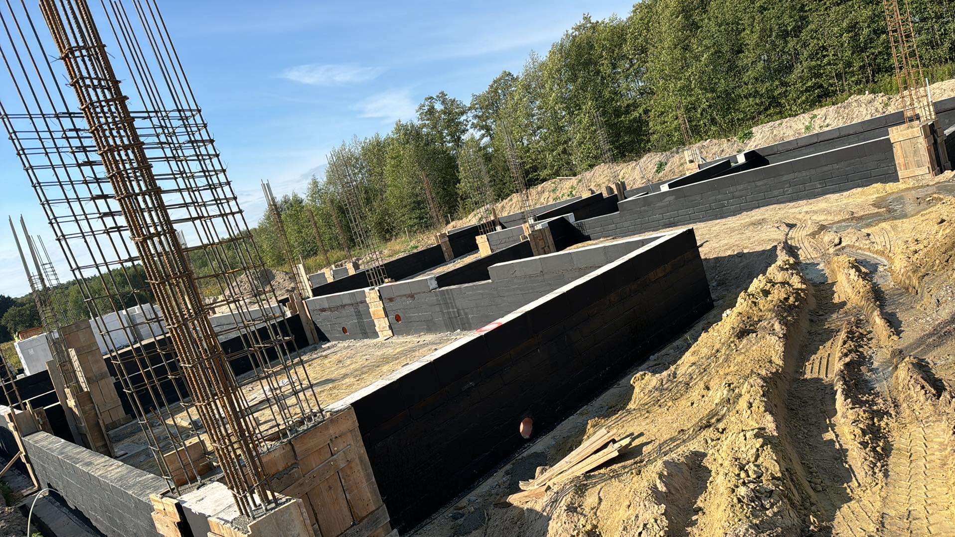 Wykopane i przygotowane fundamenty pod budynek z widocznym zbrojeniem, izolacją pionową i warstwami bloczków betonowych, otoczone ziemią i drzewami w tle.