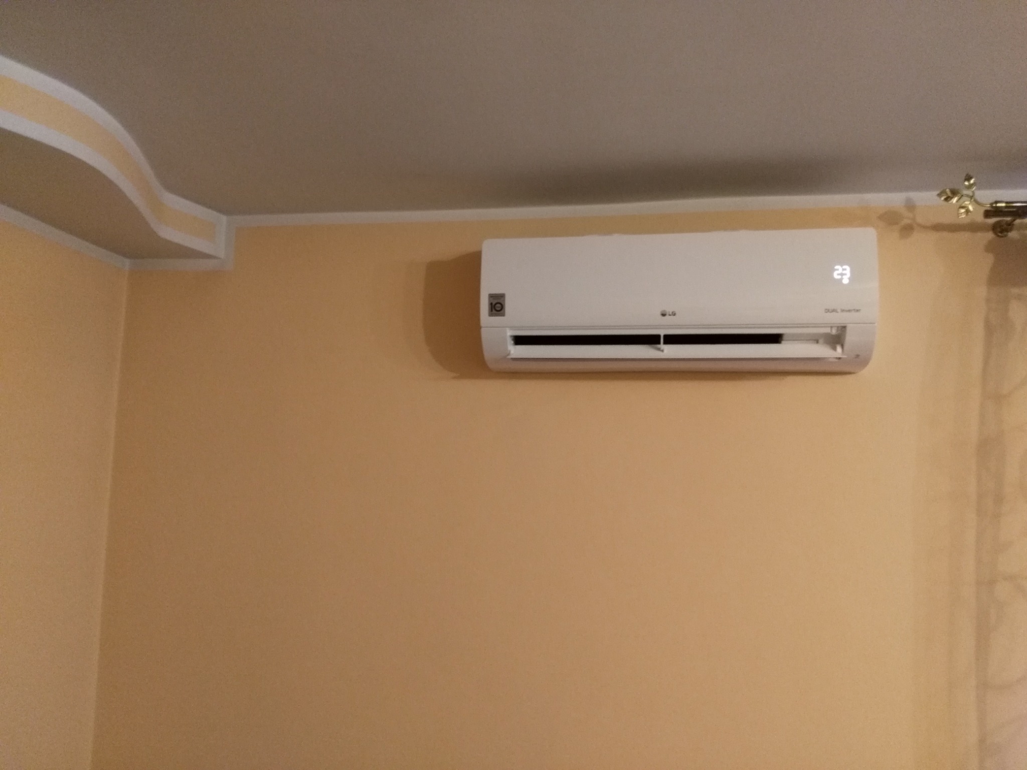 Biała jednostka wewnętrzna klimatyzacji LG zamontowana na pomarańczowej ścianie w pomieszczeniu z ozdobnym elementem sufitowym i zasłoną w tle, wyświetlająca temperaturę 23 stopnie Celsjusza.