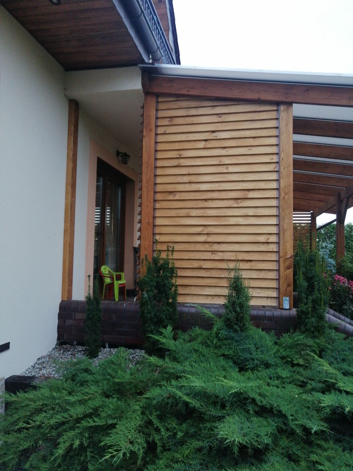 Nowoczesna pergola z drewnianą ścianą z poziomych desek, obok domu z zielonym krzesłem dziecięcym na werandzie, otoczona ozdobnymi krzewami