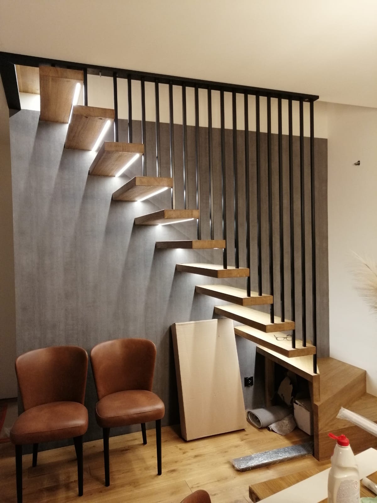 Nowoczesne schody drewniane z podświetleniem LED, czarną balustradą z pionowych prętów i dwoma brązowymi krzesłami w tle, widoczne kartony i narzędzia sugerujące montaż