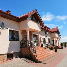 Elewacja budynku z czerwonym dachem i ceglanymi schodami prowadzącymi do wejścia, otoczona kostką brukową i zielenią.