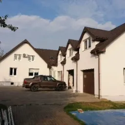 Nowo otynkowany, dwukondygnacyjny dom jednorodzinny z brązowym dachem i garażem, na posesji stoi brązowy pickup, widoczne elementy wykończeniowe.