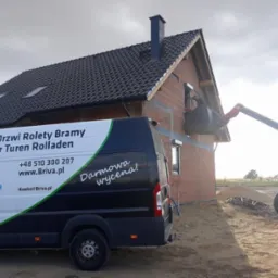 Montaż okna w nowym domu z czerwonej cegły, widoczny podnośnik teleskopowy Maniscopic i firmowy bus z napisem 'Okna Drzwi Rolety Bramy' i hasłem 'Darmowa wycena!'