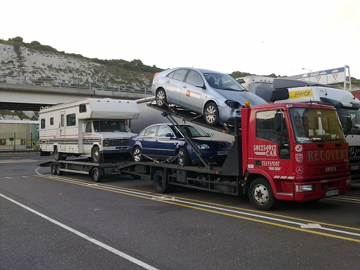 Czerwona ciężarówka z napisem 'RECOVERY' przewozi na platformie dwa samochody osobowe, w tym jeden uszkodzony z przodu, a z tyłu przyczepę z kamperem, na tle białych klifów.