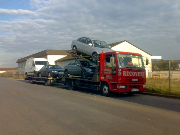 Czerwona ciężarówka z napisem 'RECOVERY' przewozi trzy samochody osobowe i furgonetkę na platformie, widok z boku na tle budynku.