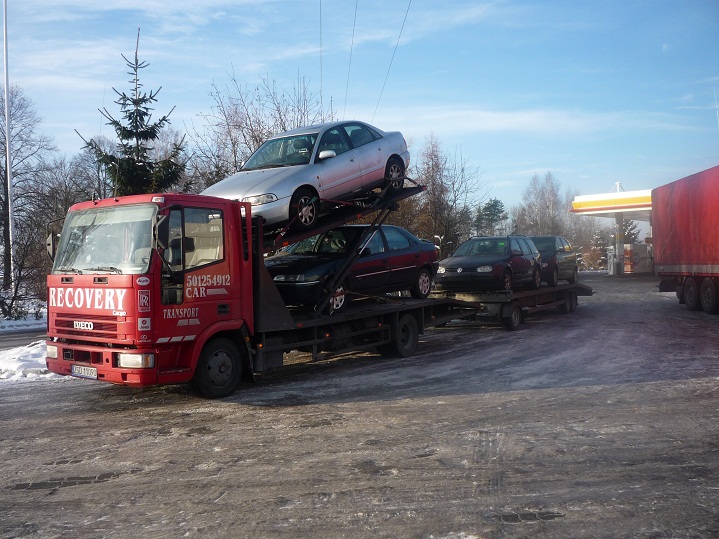 Czerwona ciężarówka z napisem RECOVERY przewozi trzy samochody osobowe na platformach, zaparkowana obok stacji benzynowej w zimowy dzień.