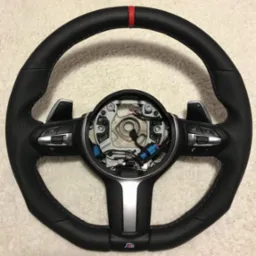 Czarna skórzana kierownica samochodowa z czerwonym paskiem na górze, niebieskimi i czerwonymi szwami, srebrnymi elementami i przyciskami, widoczne wewnętrzne okablowanie.