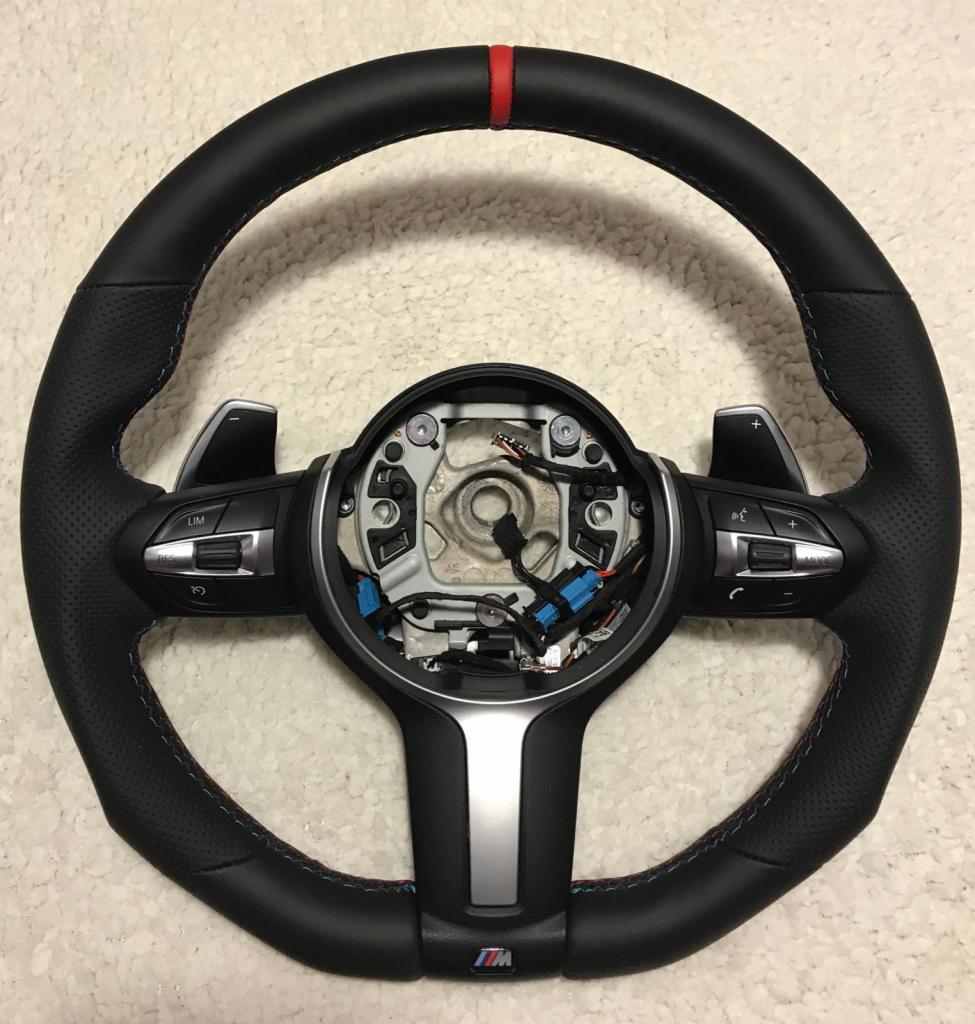 Czarna skórzana kierownica samochodowa z czerwonym paskiem na górze, niebieskimi i czerwonymi szwami, srebrnymi elementami i przyciskami, widoczne wewnętrzne okablowanie.