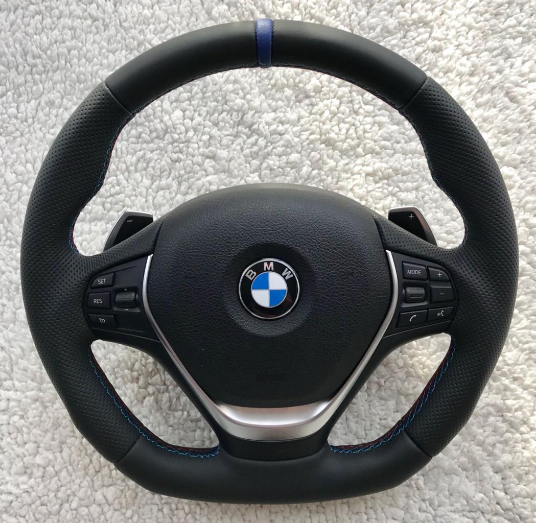 Obszyta skórzana kierownica BMW z niebieskim paskiem na górze i czerwono-niebieskimi szwami, leżąca na białym puszystym tle.