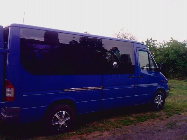 Niebieski bus Mercedes-Benz Sprinter z przyciemnianymi szybami, zaparkowany na trawiastym terenie, widoczny z boku pod kątem, z otwartymi tylnymi drzwiami.