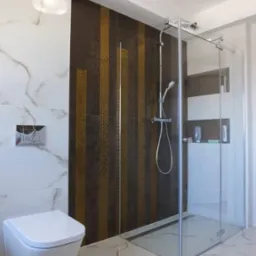 Nowoczesna łazienka z kabiną prysznicową ze szklanymi drzwiami przesuwnymi, mozaiką w odcieniach brązu i złota na ścianie prysznica, marmurowymi płytkami na podłodze i ścianach oraz podwieszaną...