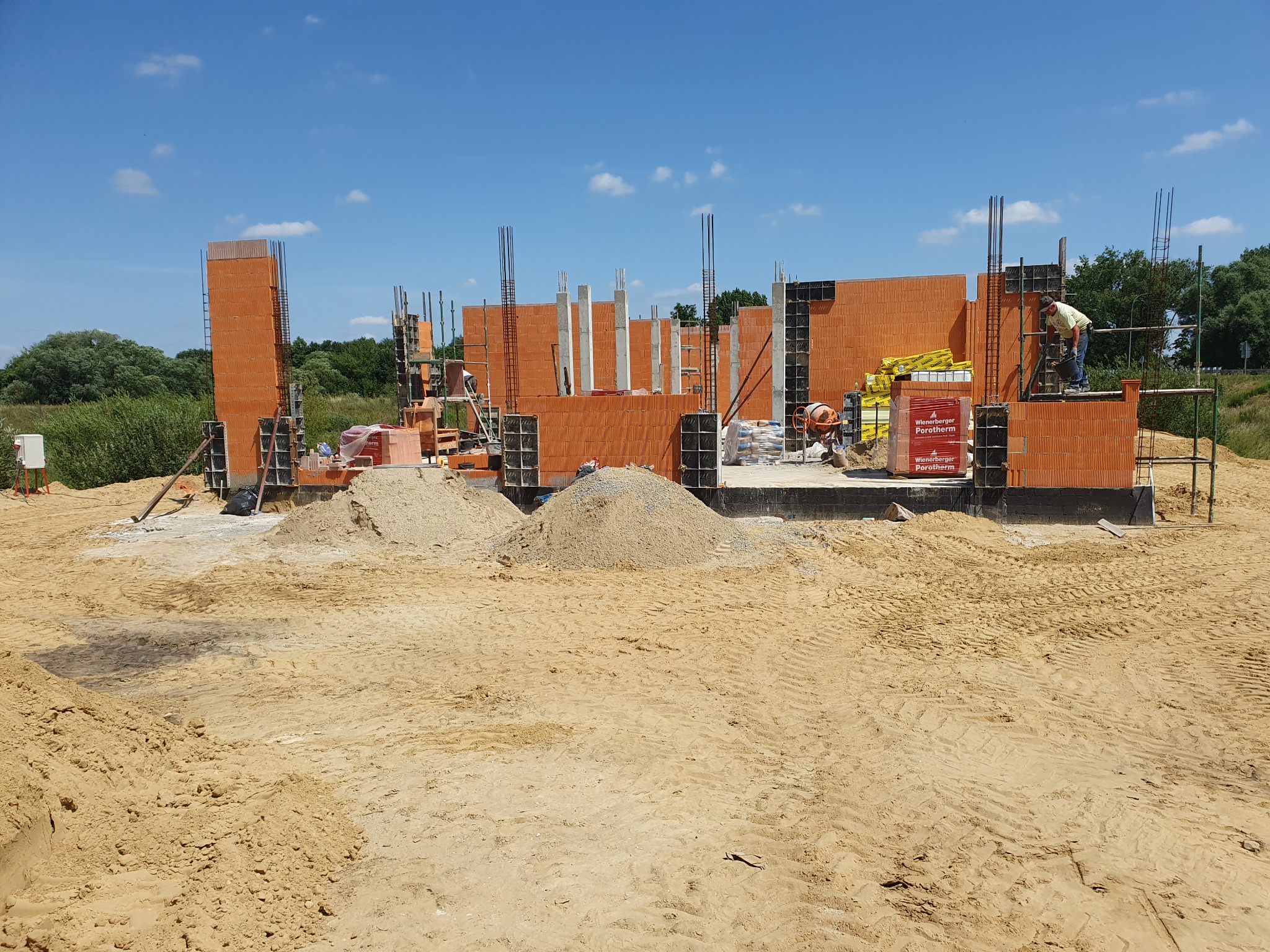 Wznoszenie ścian domu z czerwonej ceramiki, widoczne zbrojenia pionowe, palety z materiałem budowlanym i pracownik na rusztowaniu w słoneczny dzień.