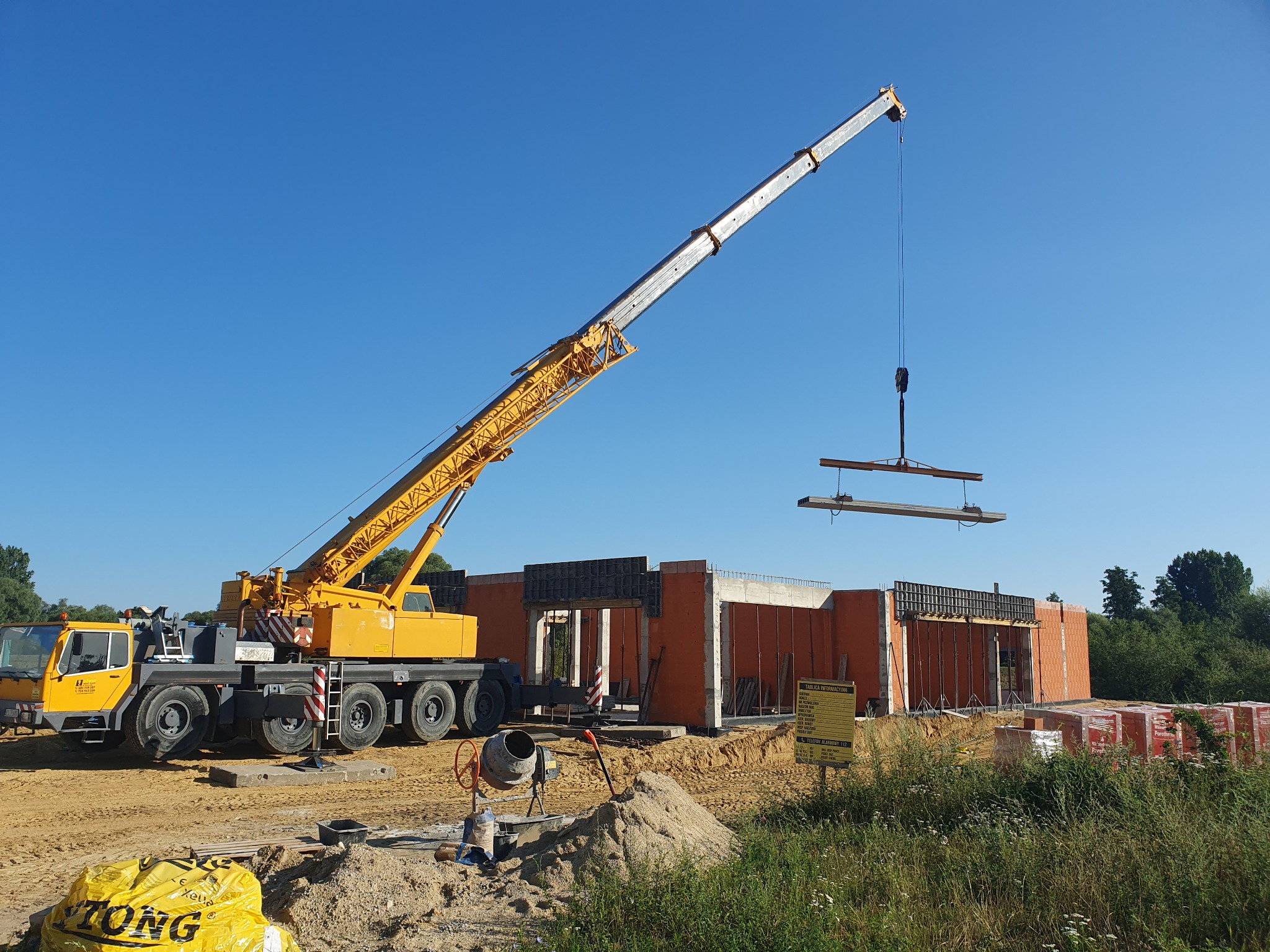 Żółty dźwig budowlany podnosi prefabrykowane elementy zbrojeniowe na placu budowy domu z czerwonej cegły. Widoczne szalunki, betonowa mieszarka i worki z zaprawą w tle błękitne niebo.