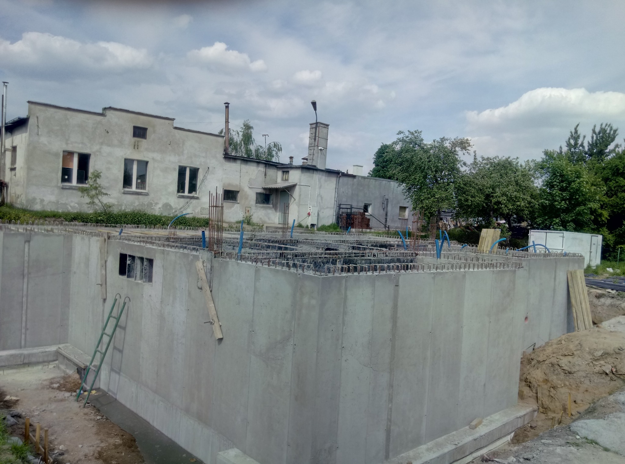 Wylane betonowe fundamenty budynku z wystającą zbrojeniem, widoczna drabina oparta o ścianę, w tle budynek gospodarczy i drzewa.