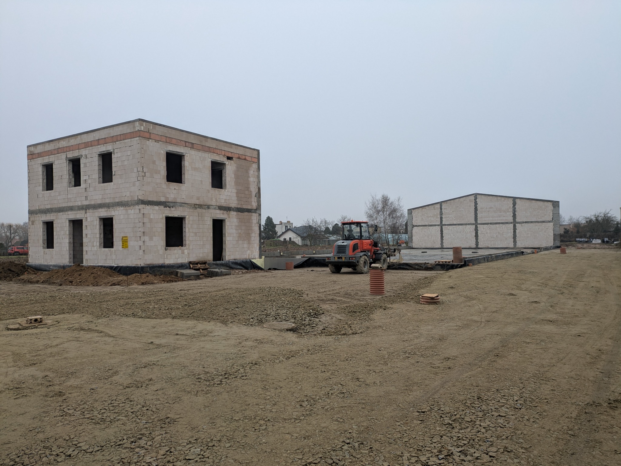 Wznoszenie dwukondygnacyjnego domu z bloczków na przygotowanym terenie, obok budowa hali, pomarańczowy traktor na placu budowy, pochmurne niebo.