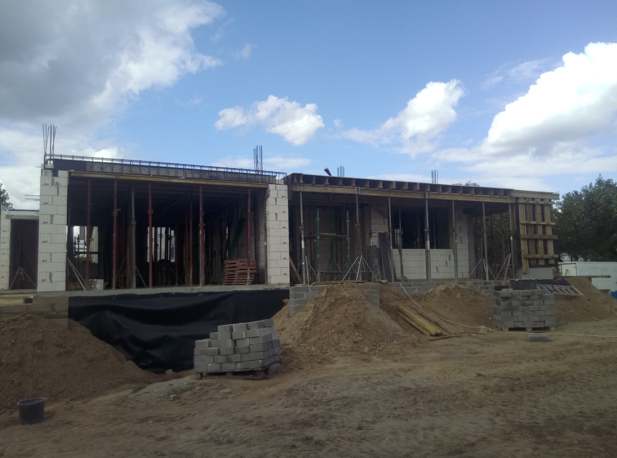 Wznoszenie ścian z bloczków betonu komórkowego, szalunki stropowe wsparte na stemplach, wystające pręty zbrojeniowe, hałdy ziemi i palety z bloczkami na placu budowy domu jednorodzinnego...