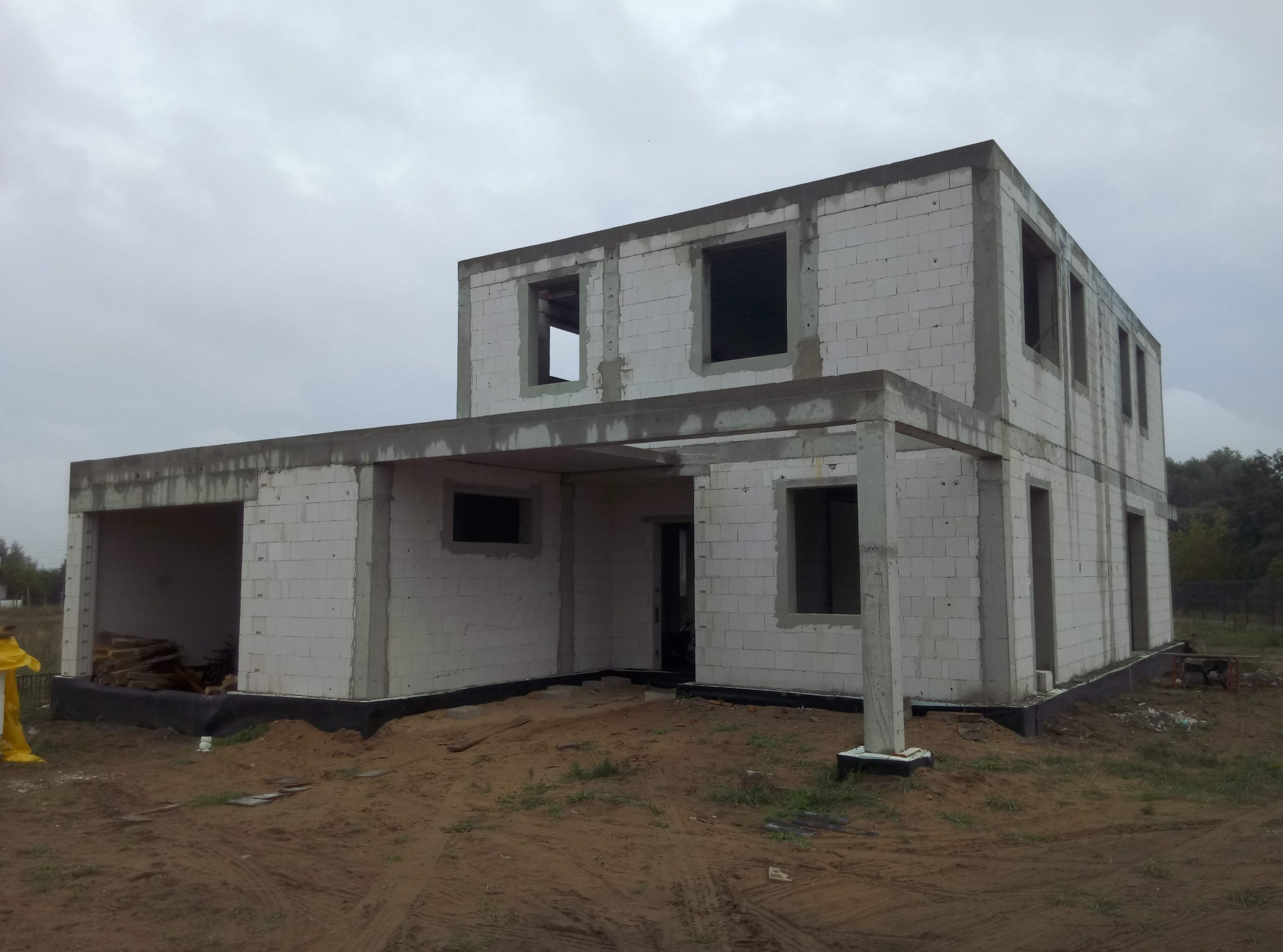 Dwukondygnacyjny budynek w stanie surowym otwartym, wzniesiony z białych bloczków, z widoczną konstrukcją żelbetową, na nierównym, piaszczystym terenie, przy pochmurnym niebie.