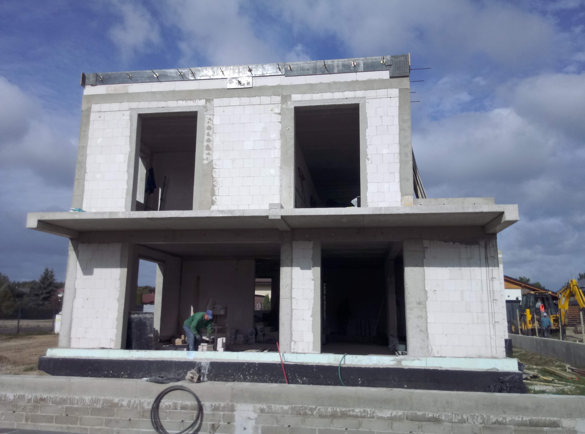 Dwukondygnacyjny dom w stanie surowym otwartym, w trakcie murowania ścian z bloczków, widoczny pracownik w zielonej kurtce, na zewnątrz koparka.