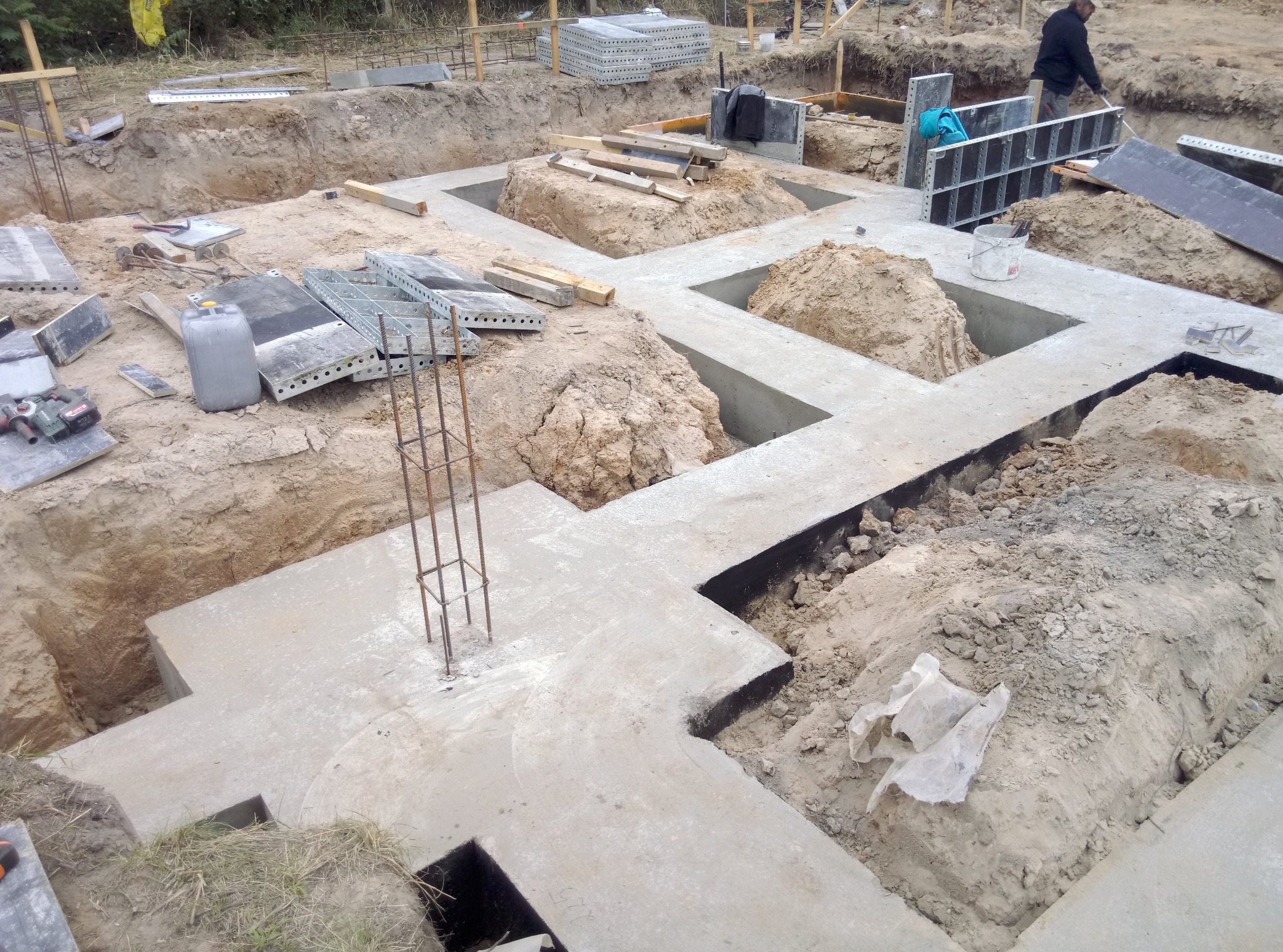 Wykop pod fundamenty z częściowo wylanym betonem, widoczne zbrojenia, szalunki i pracownik w tle.