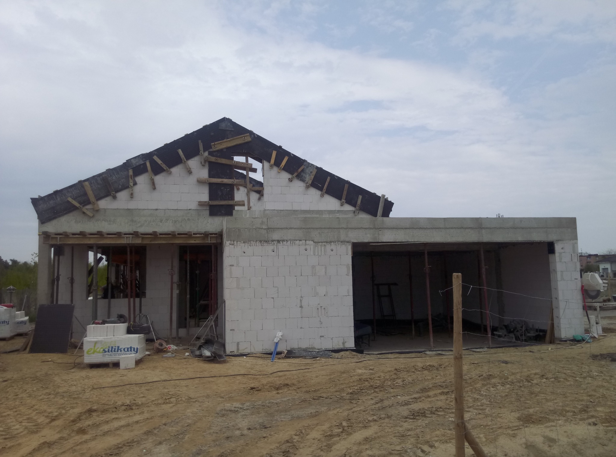 Surowy stan domu jednorodzinnego w trakcie budowy: białe bloczki silikatowe, częściowo zadaszony, widoczne drewniane elementy konstrukcyjne dachu i podtrzymujące stropy, teren wokół w stanie...