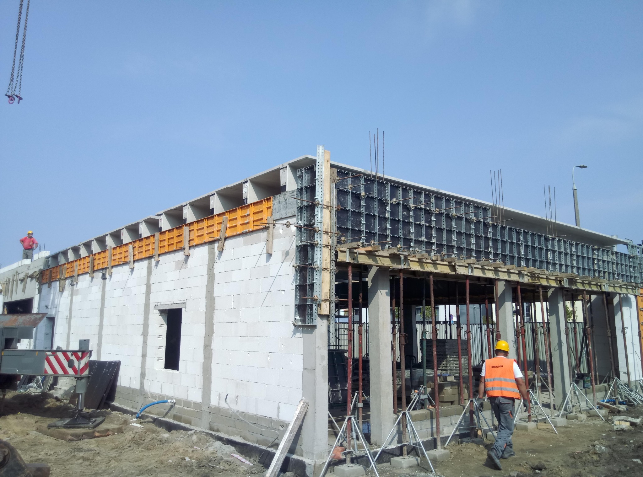 Budowa parterowego budynku z betonu komórkowego, widoczne szalunki na stropie, pracownik w kamizelce odblaskowej i żółtym kasku wchodzi do środka, dźwig z łańcuchem w tle.