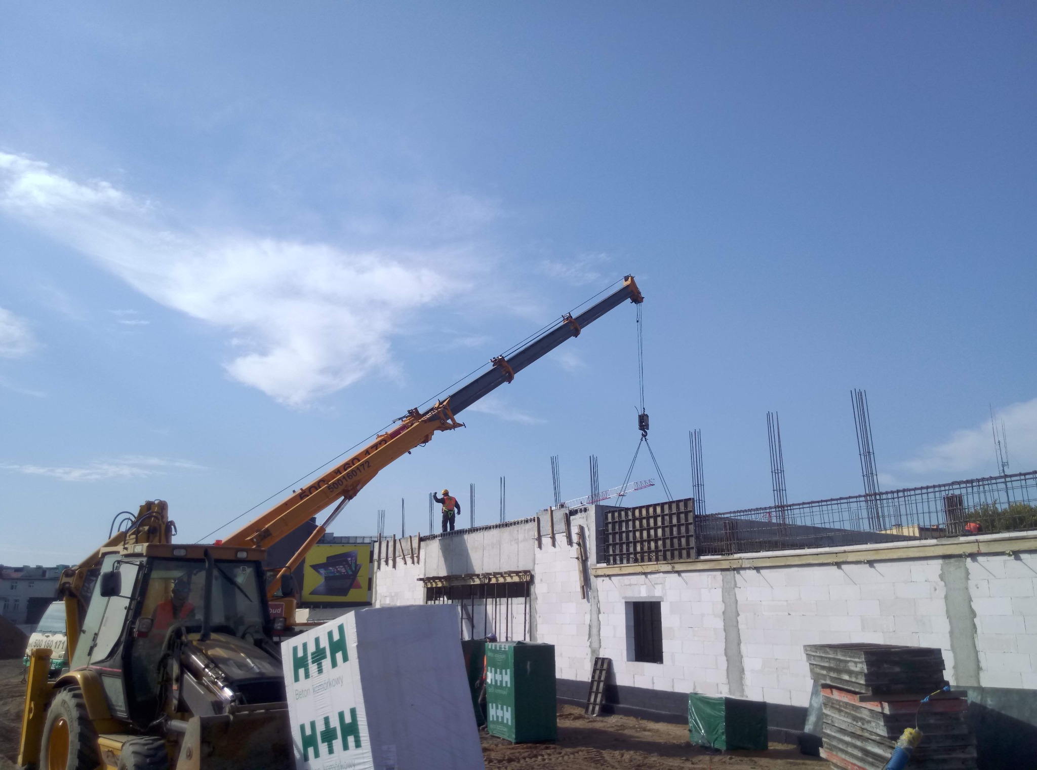 Żuraw budowlany podnosi element szalunku na budowie domu z białych bloczków, widoczny zbrojony strop i pracownik w pomarańczowym kasku na górze, na pierwszym planie palety z bloczkami H+H.
