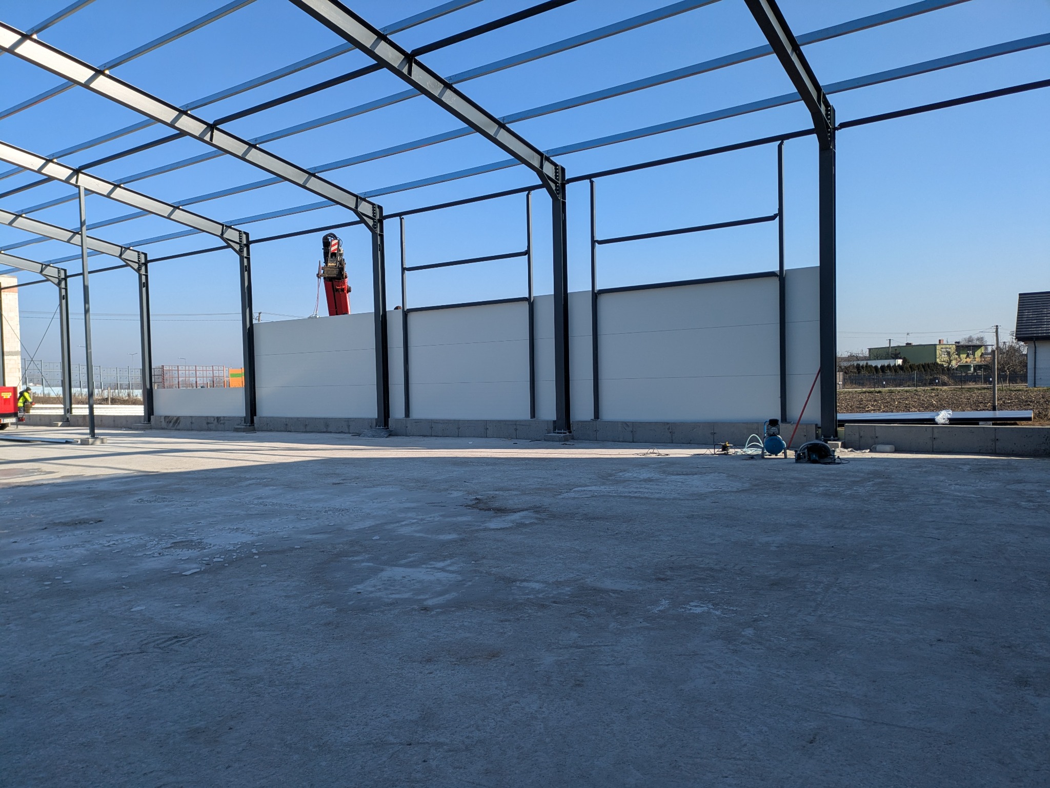 Wnętrze hali w trakcie budowy, widoczne stalowe elementy konstrukcyjne, ściany z płyt warstwowych oraz sprzęt budowlany, w tle pracownik obsługujący podnośnik.