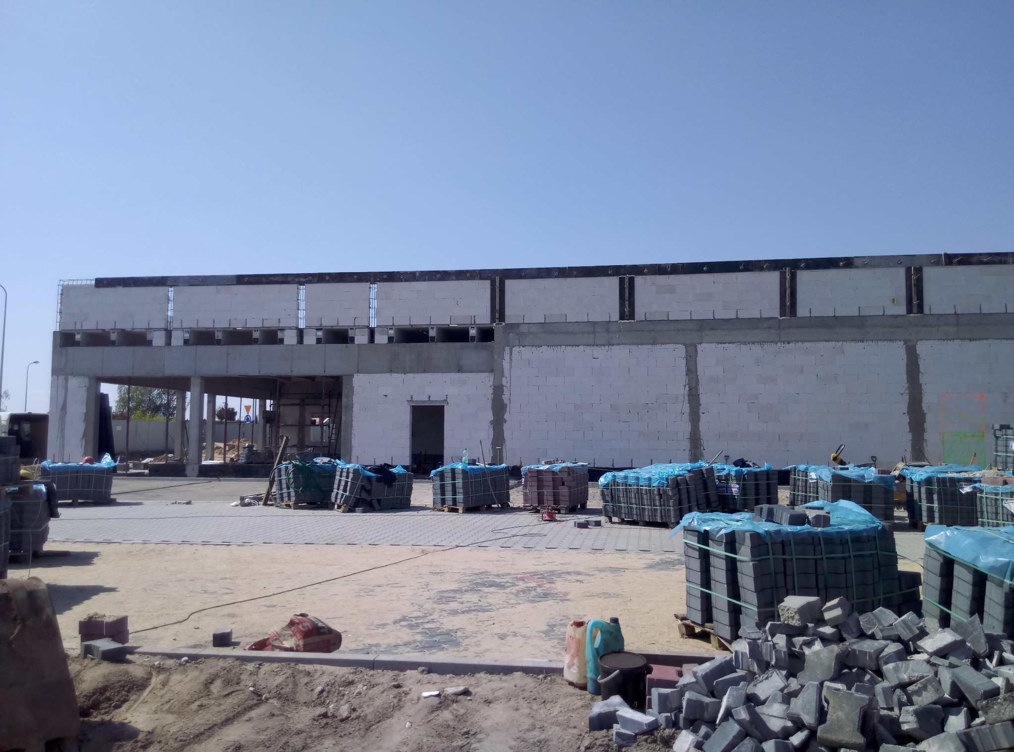 Wznoszona hala z betonu komórkowego, widoczne palety z kostką brukową, teren budowy w trakcie prac wykończeniowych.