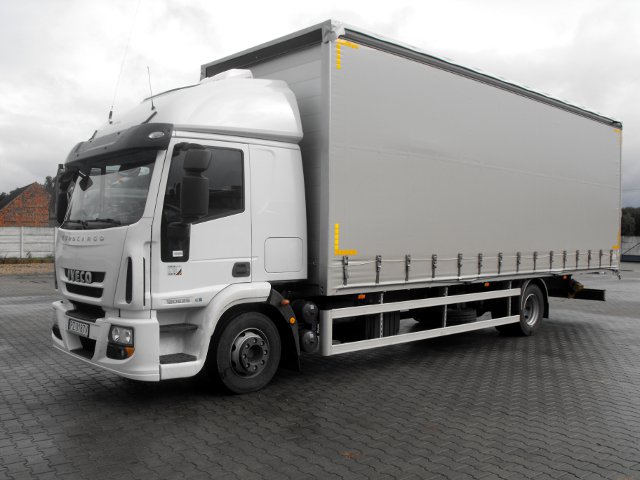 Biała ciężarówka Iveco Eurocargo z szarą plandeką na parkingu z kostki brukowej, widoczne żółte oznaczenia na plandece.