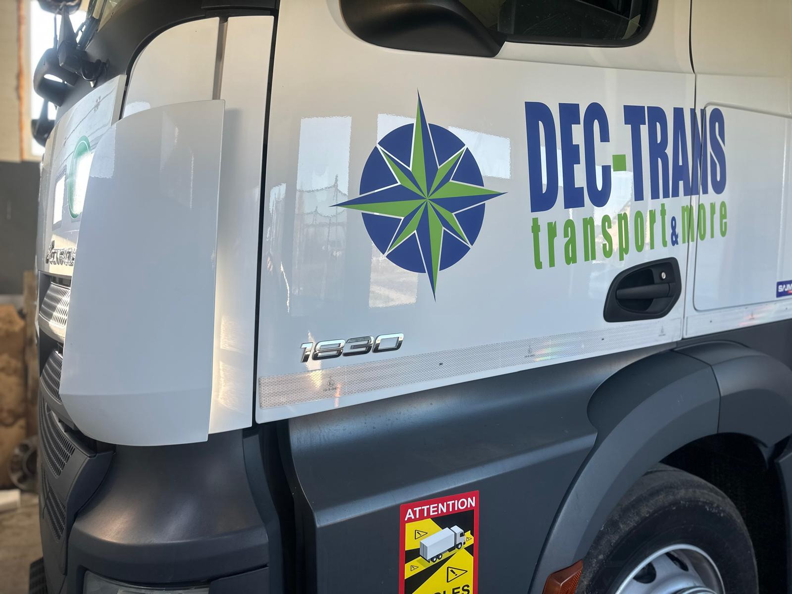 Biała kabina ciężarówki z logo firmy DEC-TRANS i symbolem kompasu. Widoczny fragment opony i naklejka ostrzegawcza. Model 1830.