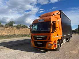 Pomarańczowa ciężarówka MAN z czarną naczepą na poboczu drogi, w tle skaliste wzgórze i błękitne niebo z chmurami. Widok z niskiej perspektywy.