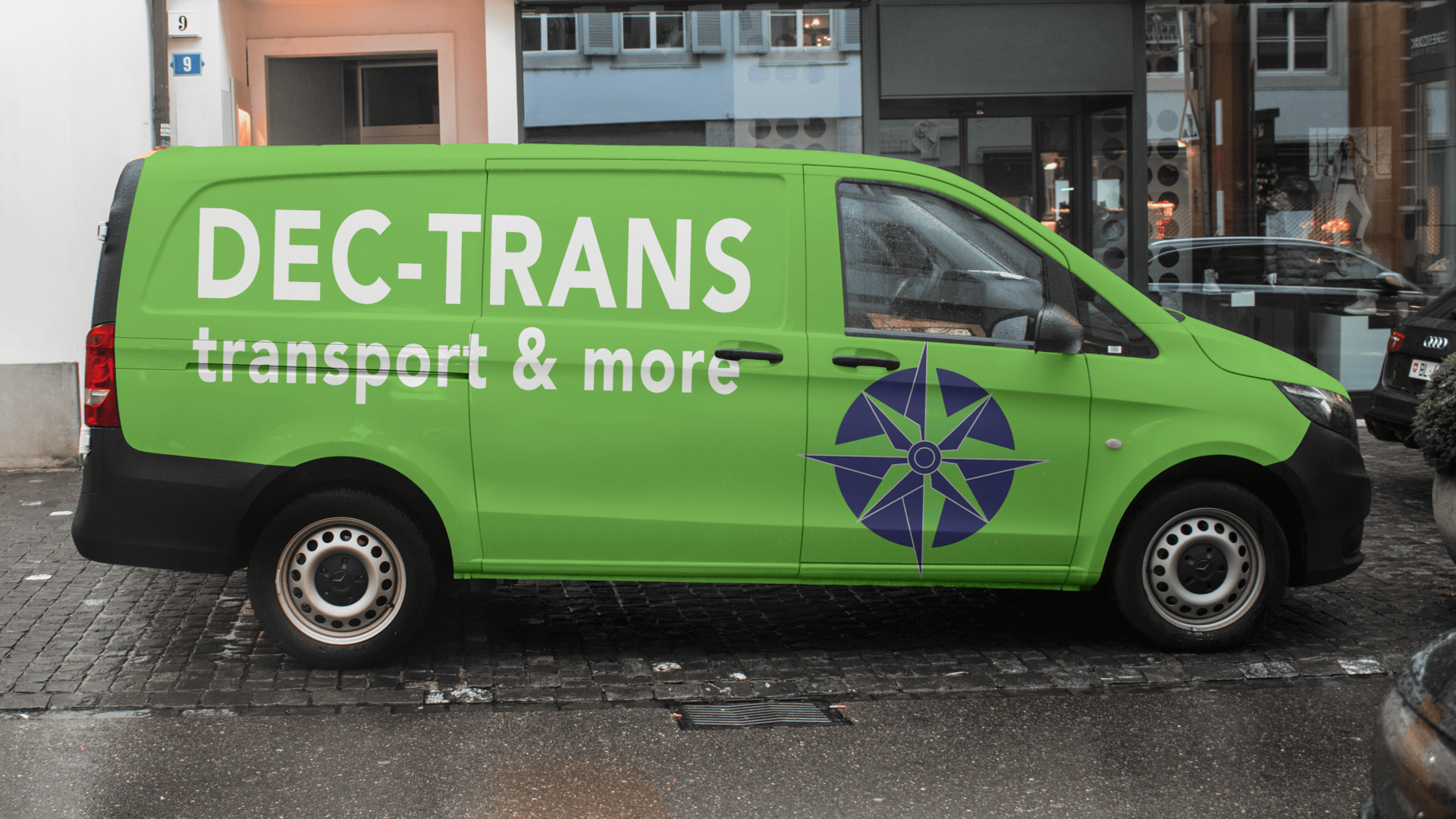 Jasnozielony van DEC-TRANS z granatowym logo kompasu na mokrej, brukowej ulicy miejskiej, widziany z boku. Napis 'transport & more' pod nazwą firmy.