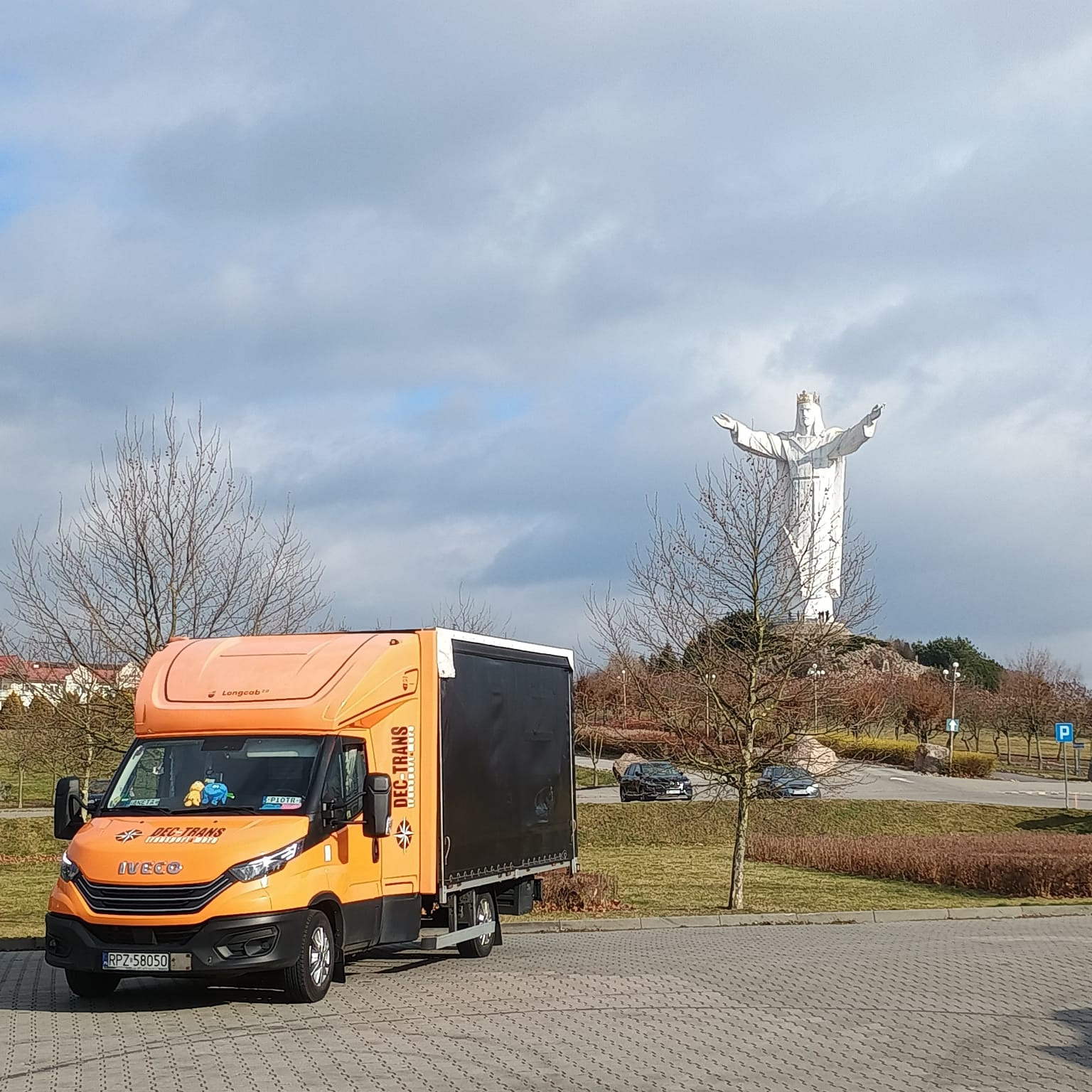 Pomarańczowy samochód dostawczy DEC-TRANS na parkingu, w tle pomnik Chrystusa Króla w Świebodzinie, pochmurne niebo.