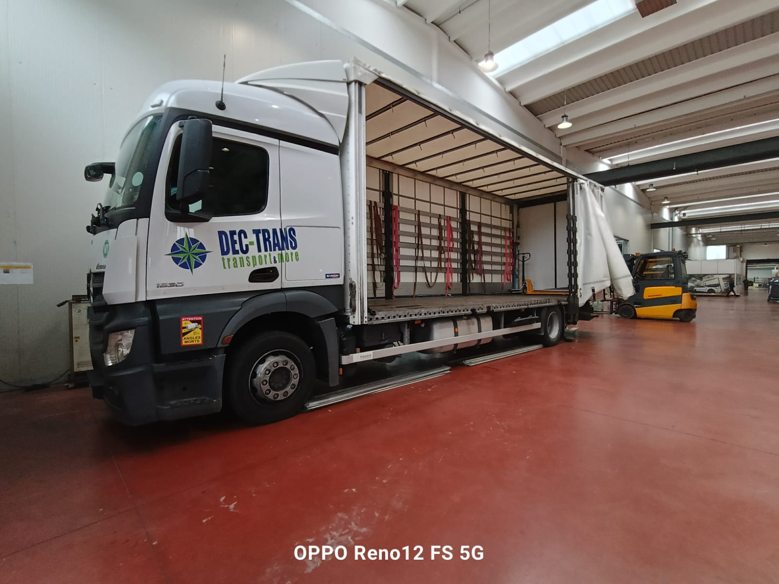 Ciężarówka z otwartą naczepą na hali magazynowej, w tle wózek widłowy. Widoczne pasy mocujące ładunek wewnątrz naczepy. Logo firmy transportowej na kabinie.