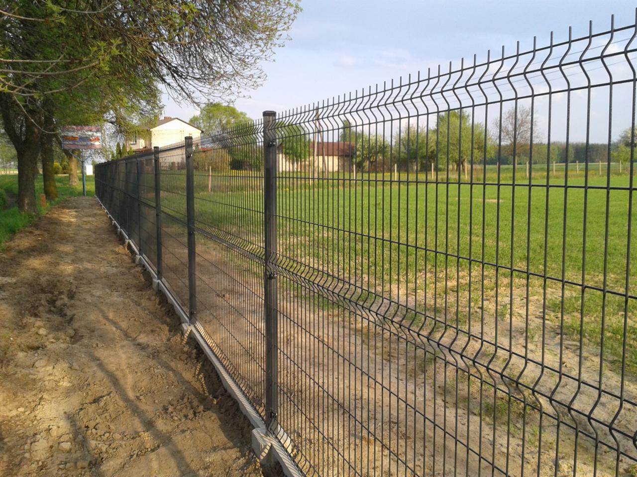 Nowoczesne ogrodzenie panelowe z metalu, widok wzdłuż linii ogrodzenia na tle zielonego pola i drzew. Słupki z czarnymi nakładkami.