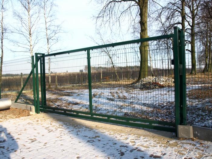 Zimowy krajobraz z zieloną, przesuwną bramą z siatki, osadzoną na betonowej podstawie. W tle drzewa i resztki śniegu na ziemi.