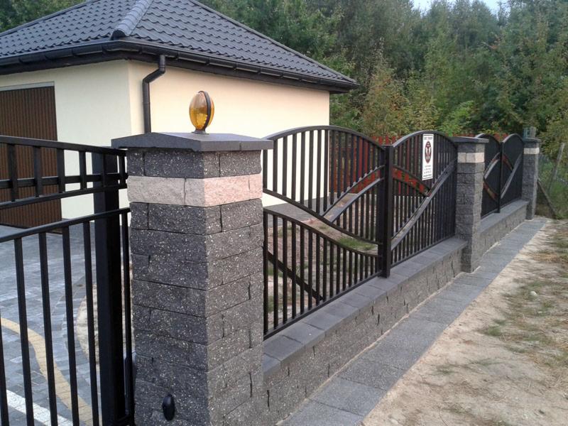 Nowoczesne ogrodzenie z czarnymi metalowymi przęsłami i słupkami z szarej kostki, zwieńczone lampą. Widok na budynek z brązowymi drzwiami i zielone drzewa w tle.