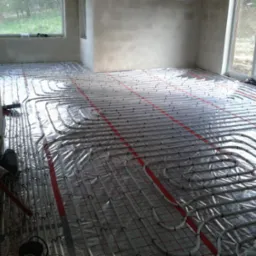 Białe rury ogrzewania podłogowego, ułożone na folii z czerwoną siatką w surowym wnętrzu. Widoczne okna i fragmenty ścian przed wykończeniem.