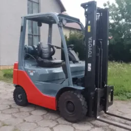 Szaro-czerwony wózek widłowy Toyota 15 na zewnątrz, na tle budynku i zieleni.