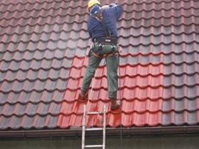 Pracownik w kasku i uprzęży na drabinie czyści dach z czerwonej dachówki, widoczna różnica między czystą a brudną powierzchnią.