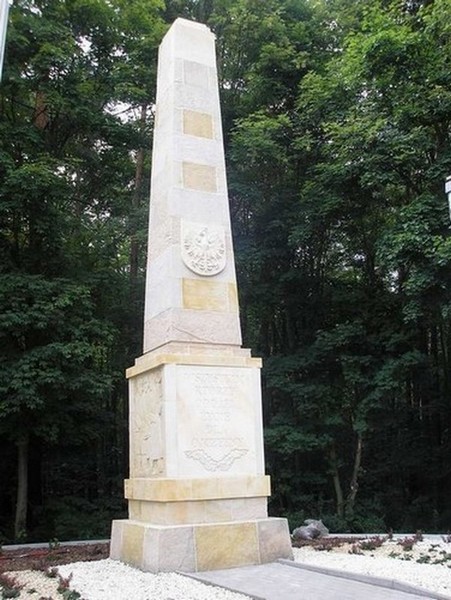 Wysoki, kamienny obelisk z polskim godłem, otoczony zielenią drzew, z wyczyszczoną powierzchnią klinkieru.