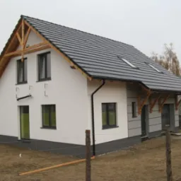 Nowoczesny dom jednorodzinny z ciemną dachówką, drewnianymi elementami wykończeniowymi i jasną elewacją. Widok z boku, częściowo ogrodzony.