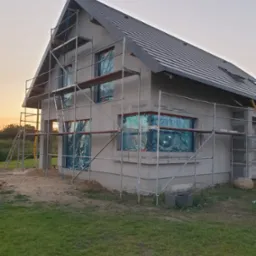 Dom jednorodzinny w trakcie prac elewacyjnych, z rusztowaniem i oknami zabezpieczonymi folią, widoczne świeżo nałożone warstwy szpachli na ścianach.