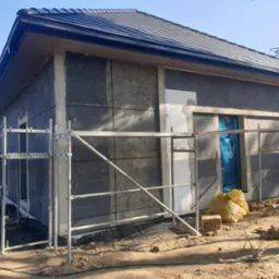 Niedokończona elewacja domu jednorodzinnego z widocznymi panelami izolacyjnymi, częściowo otynkowana, z rusztowaniem i workami budowlanymi u podstawy ściany.