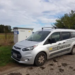 Srebrny van firmy 'Electricon' z numerem telefonu na tle rozdzielnicy SN Enea, stojący na poboczu drogi w słoneczny dzień.