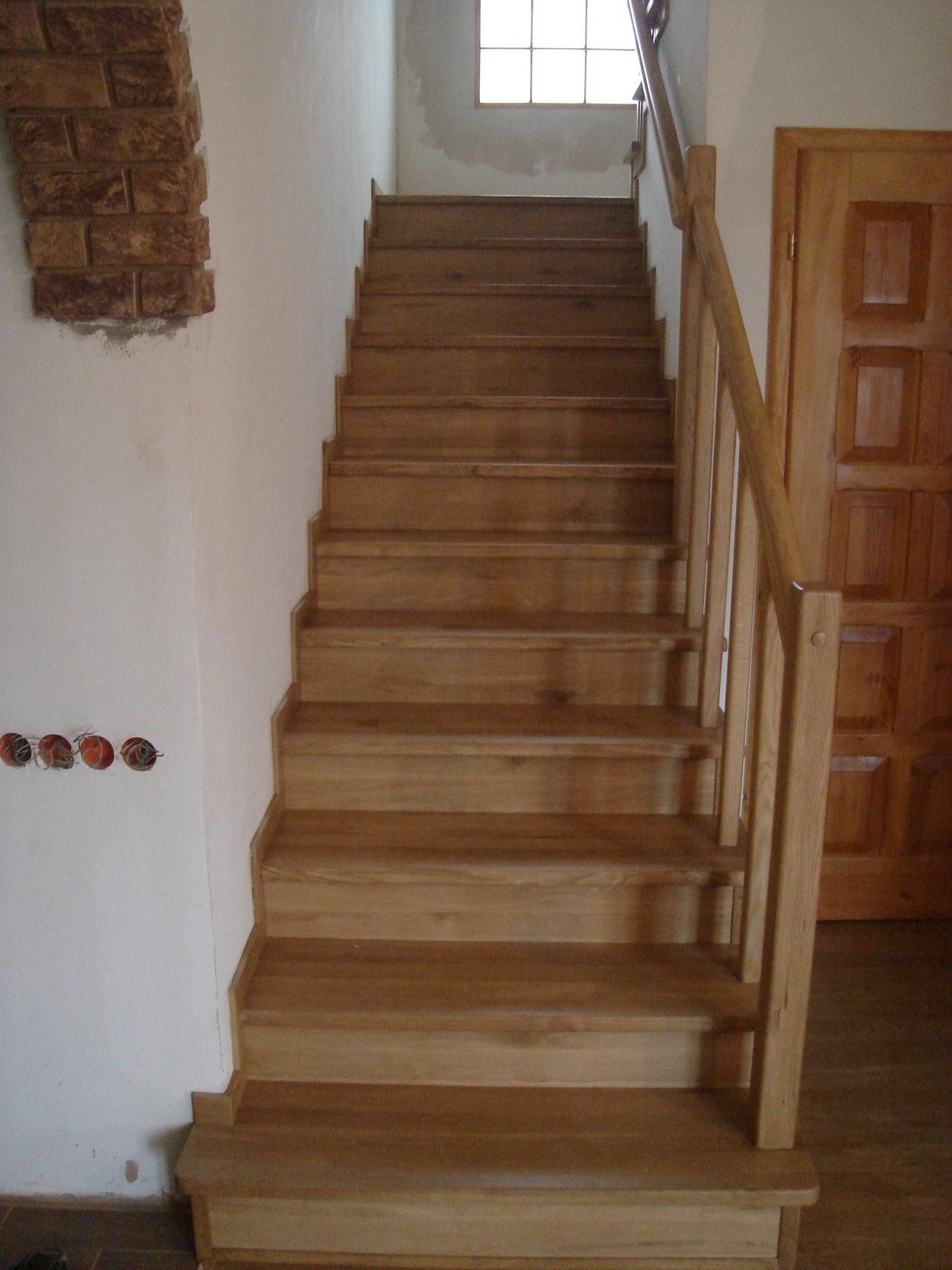 Drewniane schody z jasnego drewna prowadzące do góry, z widoczną balustradą po prawej stronie i fragmentem ściany z dekoracyjną cegłą po lewej stronie, oraz wystającymi kablami elektrycznymi.
