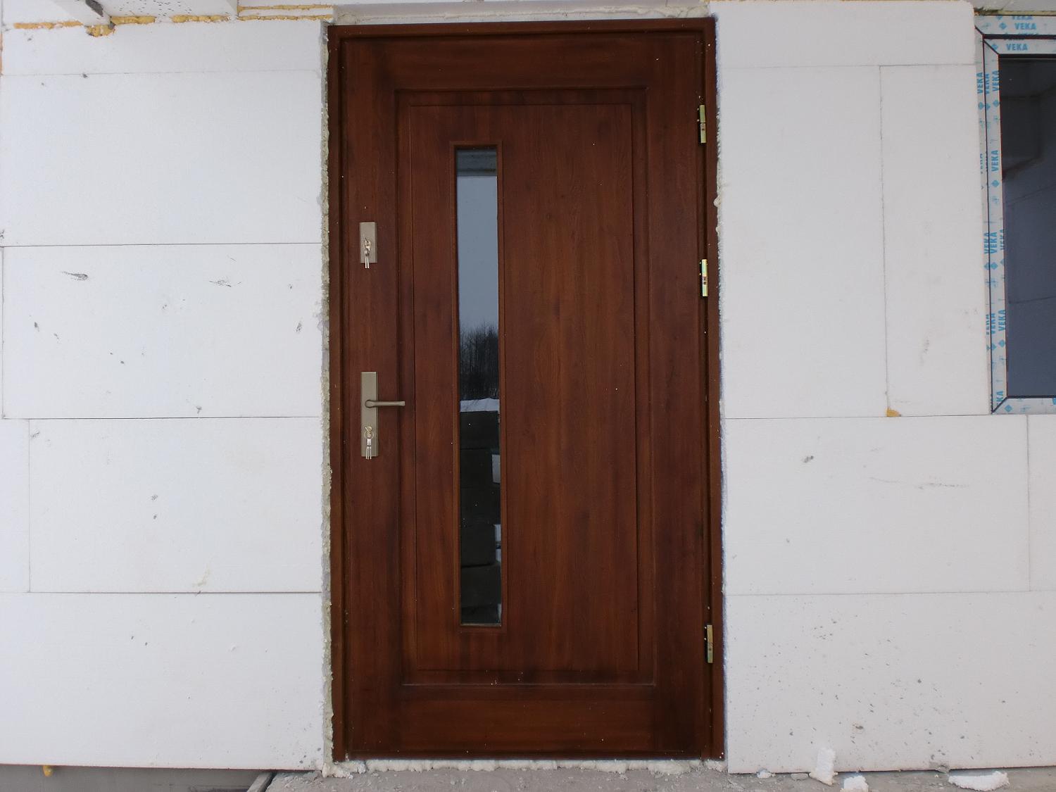 Brązowe, drewniane drzwi wejściowe z pionowym przeszkleniem, zamontowane w surowej ścianie z białych bloczków.